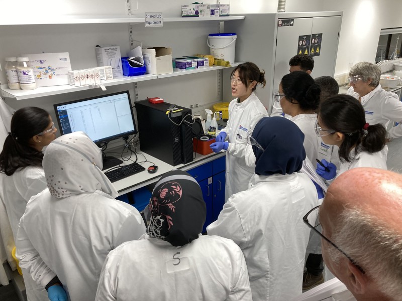 Hub members stand gathered around a computer screen displaying water sample test results, listening to a female colleague leading the workshop session